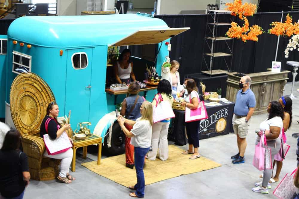 People gather at a bright turquoise food truck with a wood serving counter, receiving food and drinks. Others wait and talk nearby.