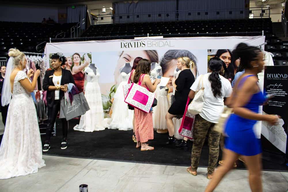 A busy bridal expo booth with a banner reading David's Bridal, surrounded by mannequins in wedding dresses and attendees.